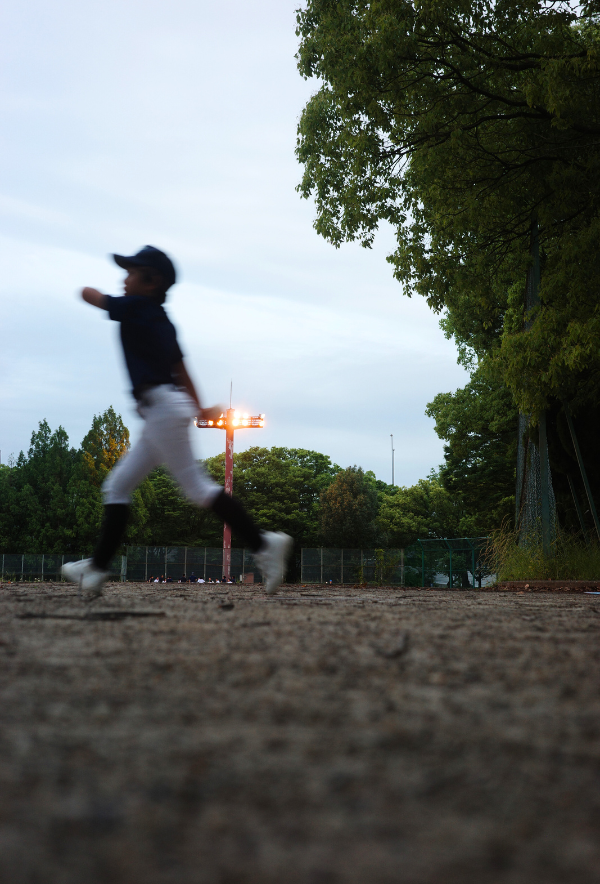 ナイター照明の下、グラウンドを駆ける少年。湿った地面と夕空のコントラストが残る、名古屋・楠公園の一瞬。