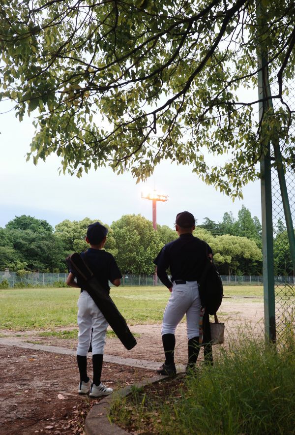 野球道具を抱えたふたりの少年が、ナイター照明に照らされるグラウンドを静かに見つめている、雨上がりの夕暮れ。
