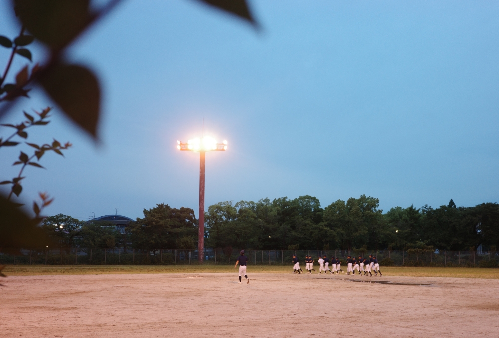 グラウンドを横切る子どもたちの列。照明と夕空が淡く重なり合う、名古屋・楠公園のナイター前の時間。