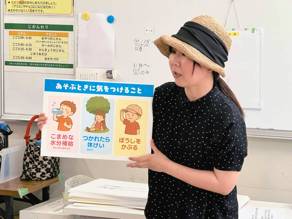 教室で帽子をかぶった女性が「こまめな水分補給」「つかれたら休けい」「ぼうしをかぶる」と書かれた熱中症対策のポスターを持ち、子どもたちに説明している様子。