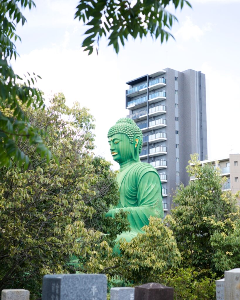 名古屋市千種区・桃厳寺の名古屋大仏。住宅街のマンションと墓地を背景に、緑の大仏が木々の間から姿を現す風景