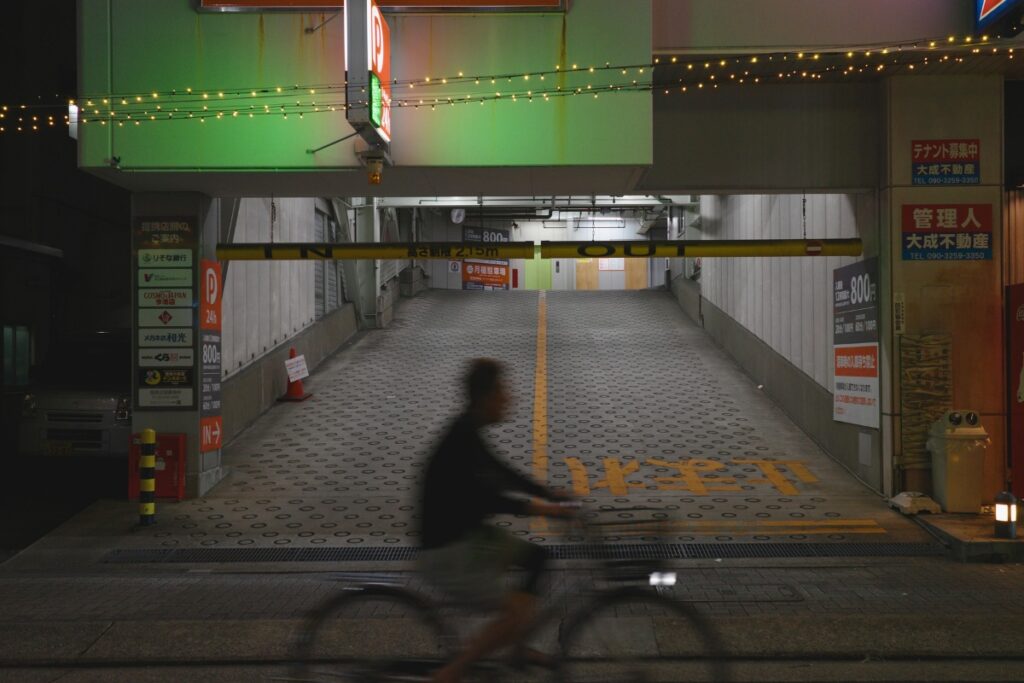 名古屋・今池の夜。駐車場の前を自転車で走る人のシルエット。せんべろ元気がある今池の雑多な街並みを象徴する光景