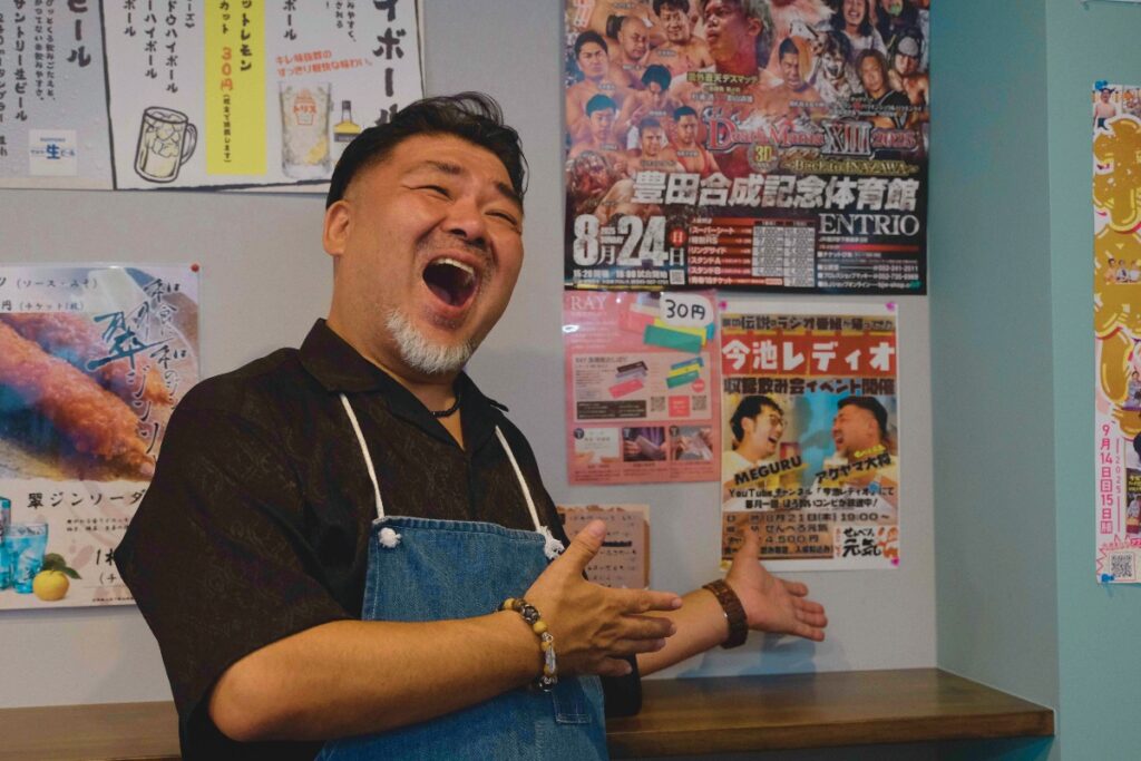 名古屋・今池の居酒屋せんべろ元気店内で、今池レディオのポスターを背景に笑顔を見せる店主・明山聡一郎