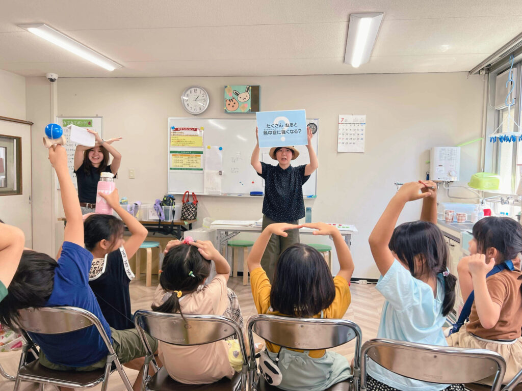 教室でスタッフが「たくさんねると熱中症に強くなる？」と書かれたボードを掲げ、子どもたちが手や体で〇や✕のポーズをとりながらクイズに答えている様子。