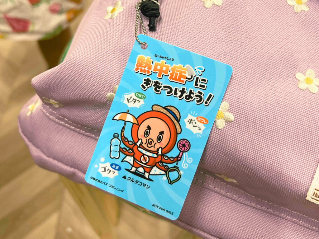 紫色の花柄リュックに付けられた、熱中症予防を呼びかける「熱中症に気をつけよう！」と書かれたカード。キャラクターのタコ「クルタコマン」が水分補給や帽子をかぶるなどの予防行動を呼びかけている。