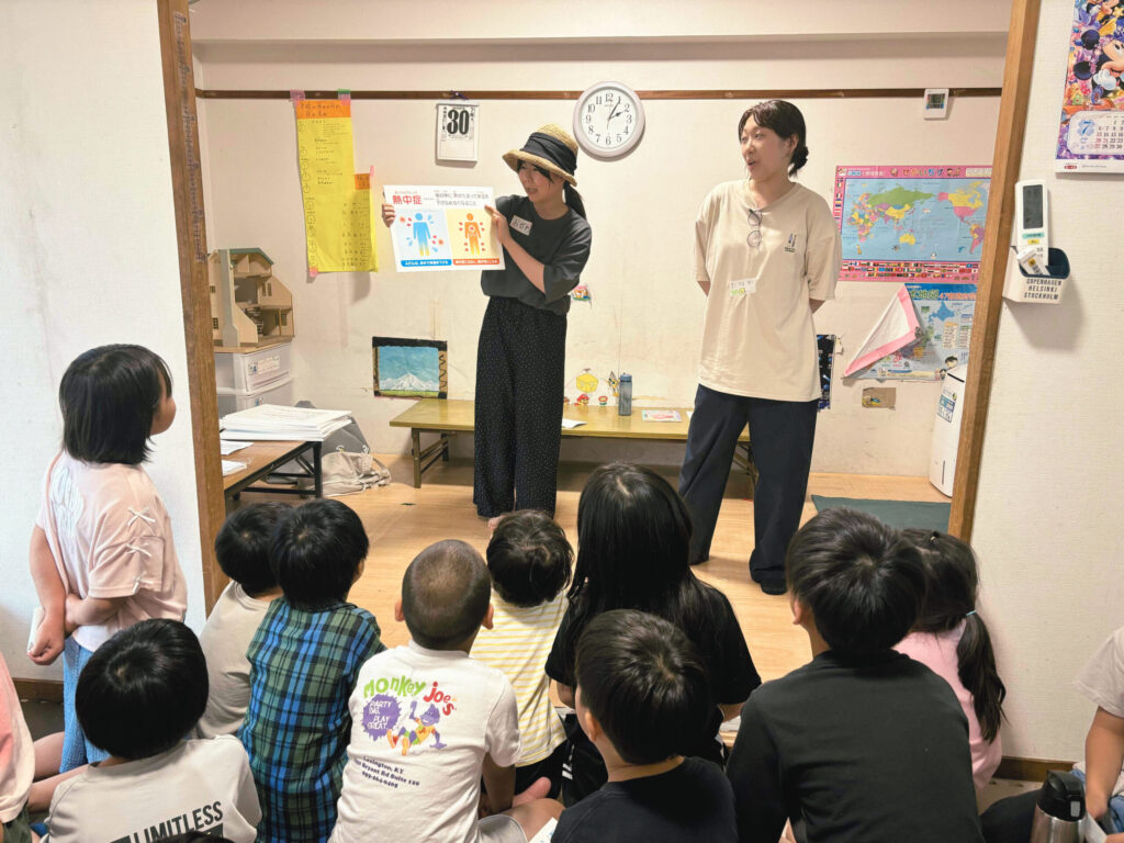 室内で帽子をかぶった女性が熱中症予防のポスターを持ち、隣に立つスタッフと一緒に子どもたちへ説明している様子。床に座った複数の子どもたちが前を向き、真剣に話を聞いている。