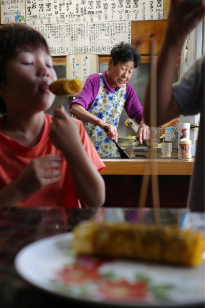 名古屋市北区上飯田の駄菓子屋「つねかわ」で、スナックロールを焼くお母さんと、それを楽しそうに頬張る子どもたち。店内には手書きのメニューが並び、昭和の空気が漂う。
