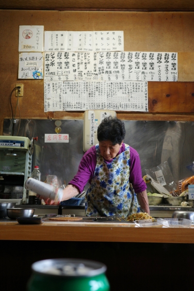 駄菓子屋「つねかわ」の店内で鉄板を前に調理するお母さん。壁には長年の手書きメニューが残り、時代の積み重ねを感じる。