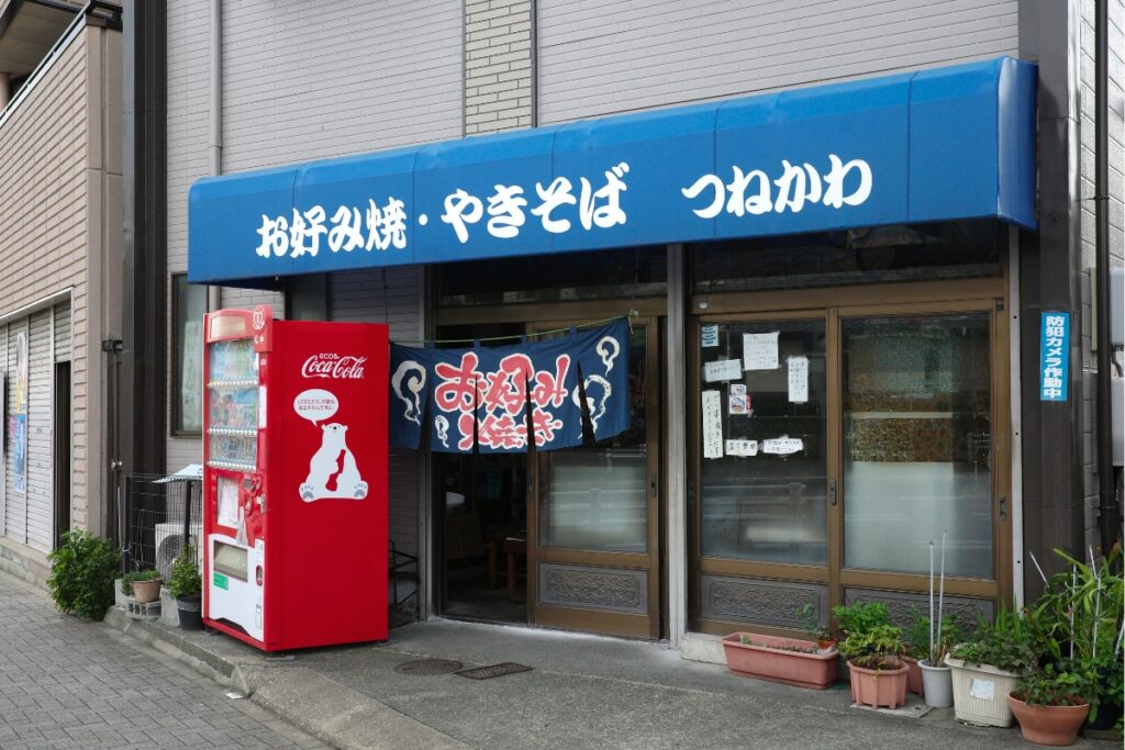 青いテントに「お好み焼・やきそば つねかわ」と書かれた店舗外観。赤い自販機とのコントラストが印象的な、名古屋市北区上飯田の駄菓子屋・鉄板焼き屋。