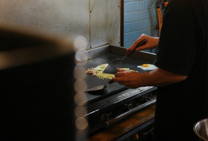 鉄板の上で焼きそばを調理する様子。両手にヘラを持ち、麺と具材、目玉焼きを丁寧に焼く姿。金山南の「KING焼きそば」店内での一場面。