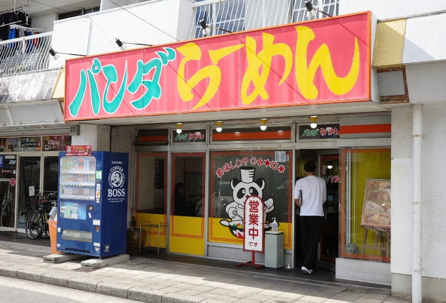 団地の下に並ぶ店舗の中で、赤と黄色の派手な看板が目立つパンダラーメンの外観。昔ながらの雰囲気と強い存在感が伝わる。