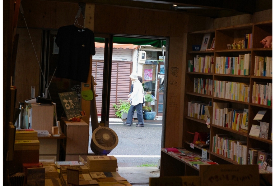 「ブタコヤブックスの店内から外を眺める視点。開いた扉の向こうを、ゆっくり歩く人の姿が通り過ぎていく。笠寺の日常が静かに流れる一瞬。