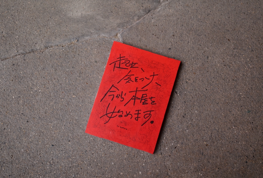 赤い表紙のZINE『起立、気をつけ、今から本屋を始めます。』が、笠寺の路面にそっと置かれている様子。