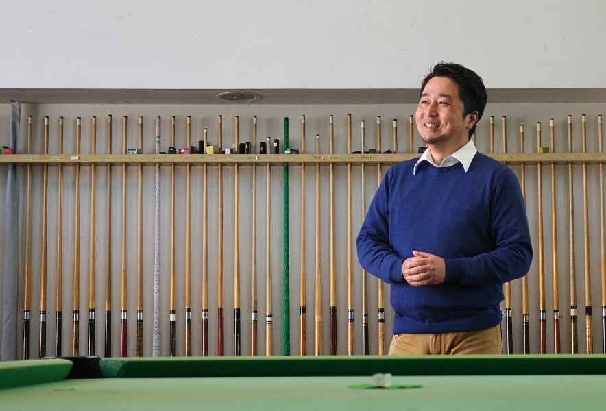 名古屋市昭和区・曙通りのビリヤード場「友楽」。キューが並ぶ壁を背に、笑顔の店主が静かに立つ。