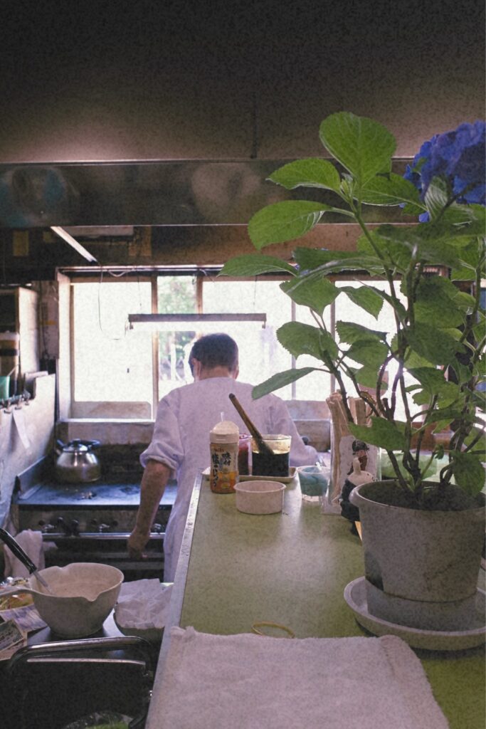 カウンター越しに見える板鼻氷店の店内。鉄板に向かうお母さんの背中と、長年積み重ねられた日常の風景。