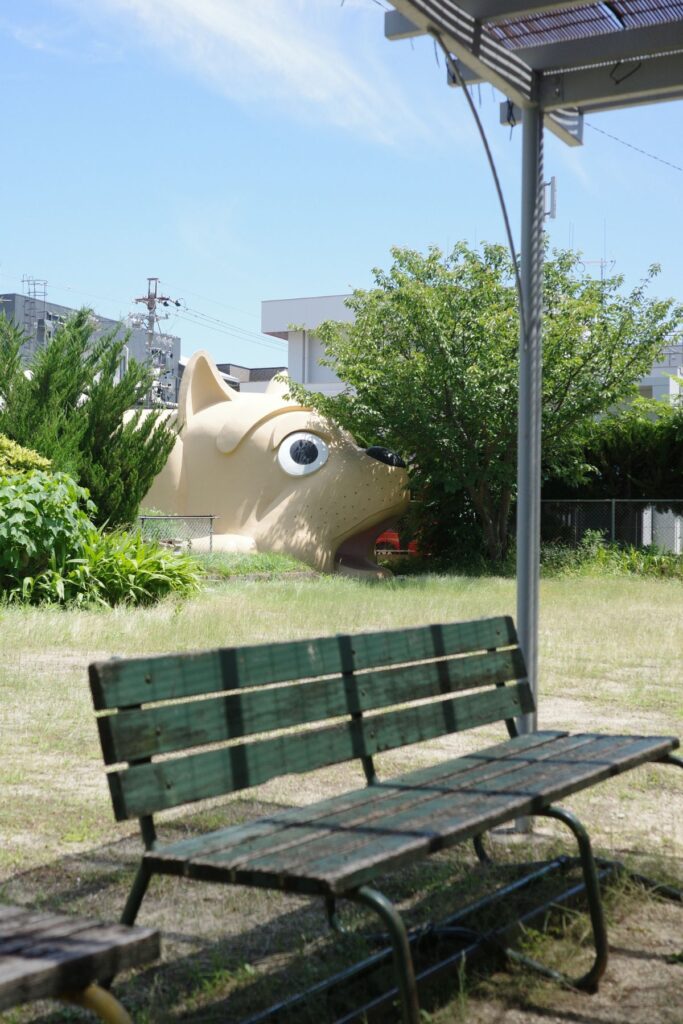 ベンチ越しに見える巨大な犬型消防団詰所。若葉児童遊園地の日常風景。