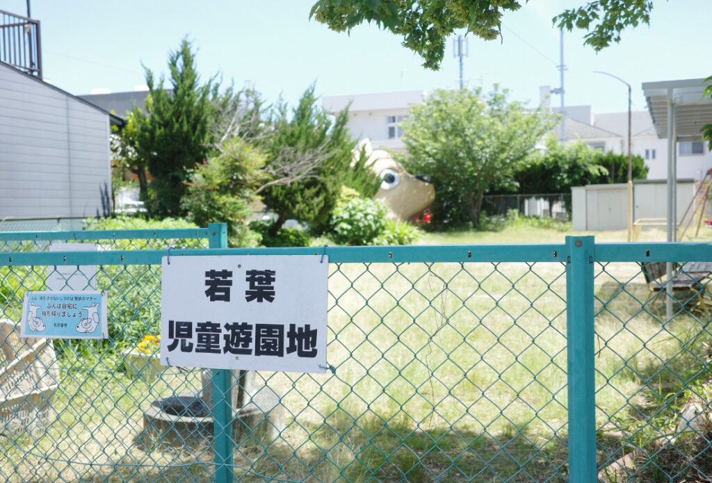 若葉児童遊園地の看板。その奥には木々に隠れるように巨大な犬の姿が見える。