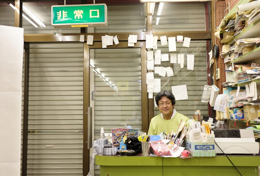 非常口の案内板の下、メモが貼られたシャッター前に座る店主