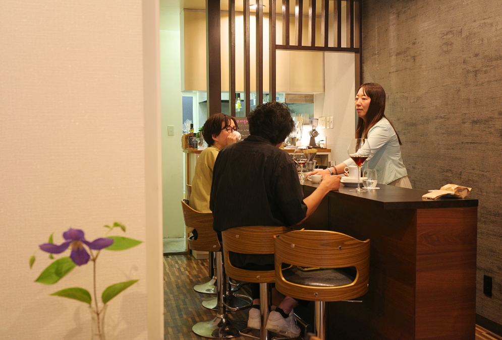 「Grains de café」のカウンターで、お客さんと会話する店主・三冨由杏子さん。5席の小さな店内でコーヒーを楽しむ様子。