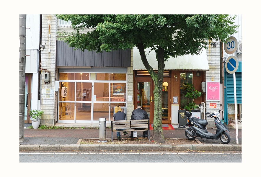 名古屋市南区・内田橋商店街。ミュージックバー「Quest Luv」の前のベンチでくつろぐ2人と、街路樹のある通り。