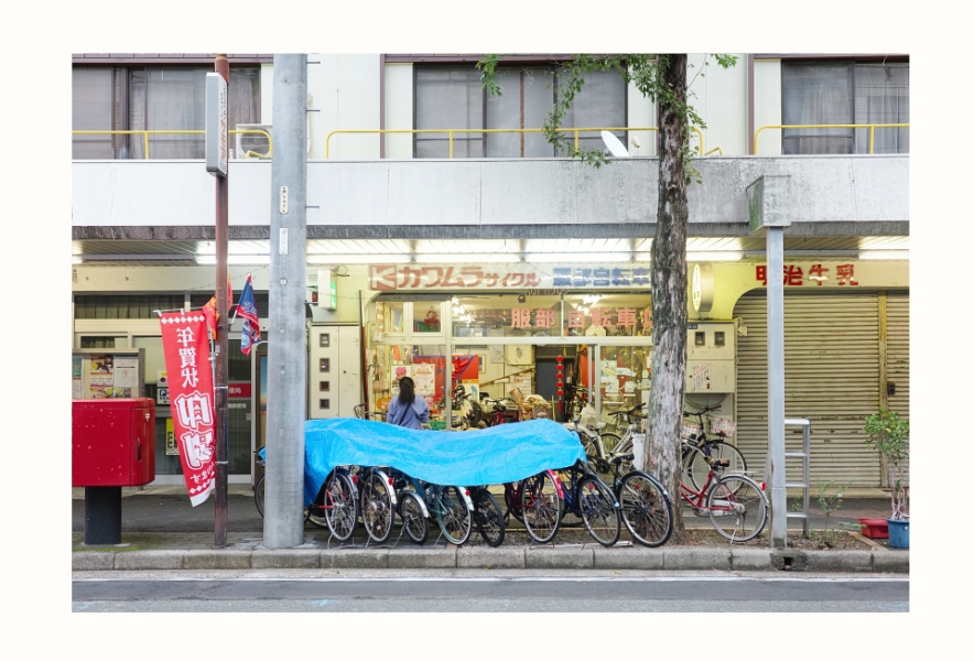 名古屋市南区・内田橋商店街の街並み。昔ながらの建物の1階に自転車店があり、店先には何台もの自転車が並ぶ。青いシートや色褪せた看板、赤い郵便ポストが、商店街の日常を映し出している。