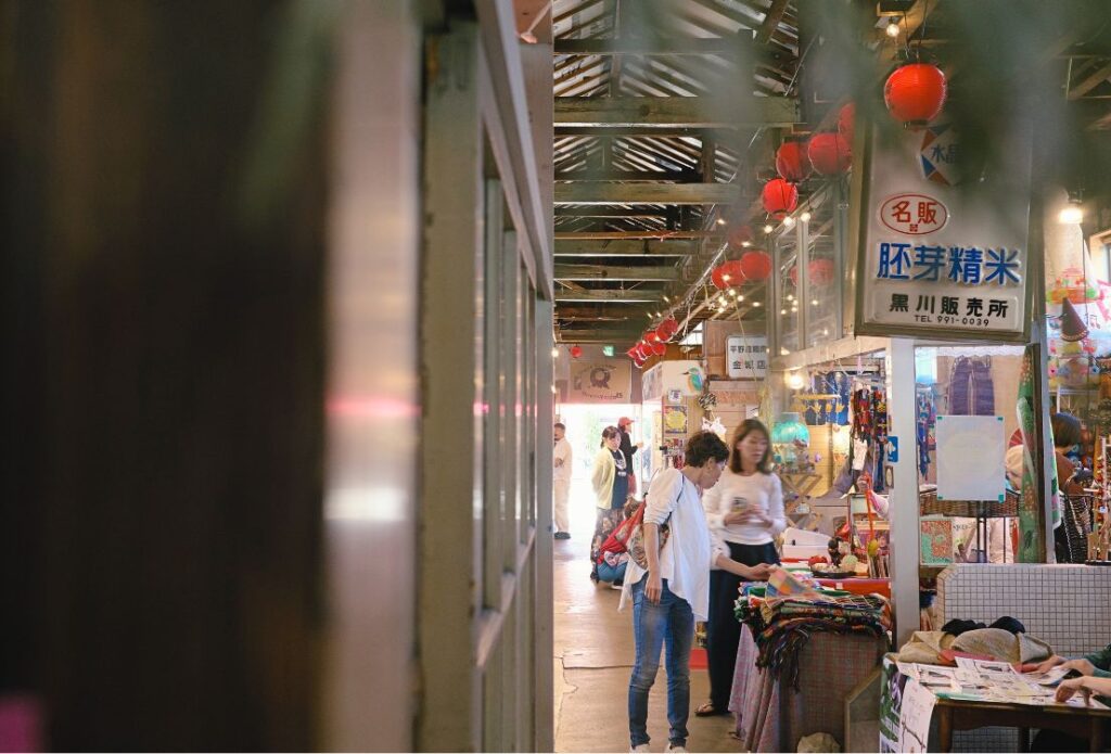 金城市場の通路。昔の「胚芽精米」の看板と赤い提灯の下で、朝市の買い物を楽しむ人たち