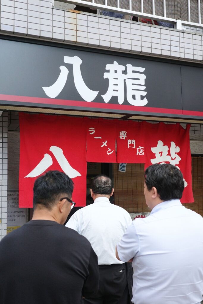 ラーメン専門店「八龍」千代田店の赤い暖簾の前で、昼どきに並ぶ人たち