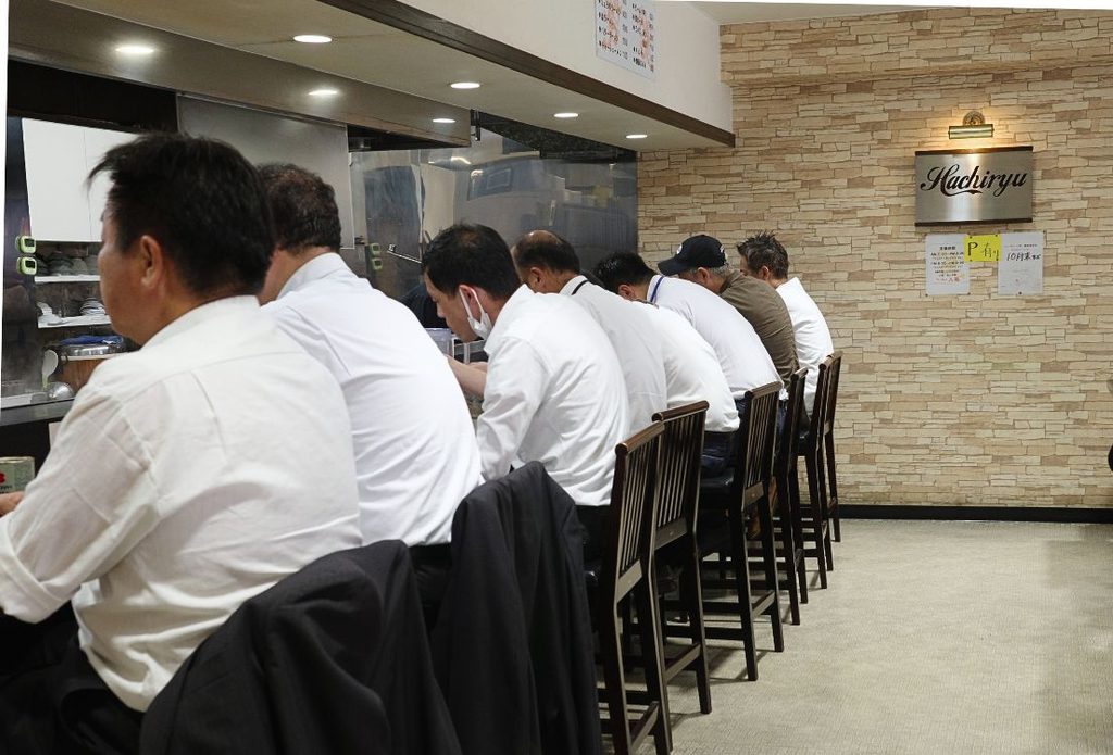 名古屋・八龍 千代田店のカウンターに並び、昼休みに味噌ラーメンを食べる白いワイシャツのサラリーマンたち