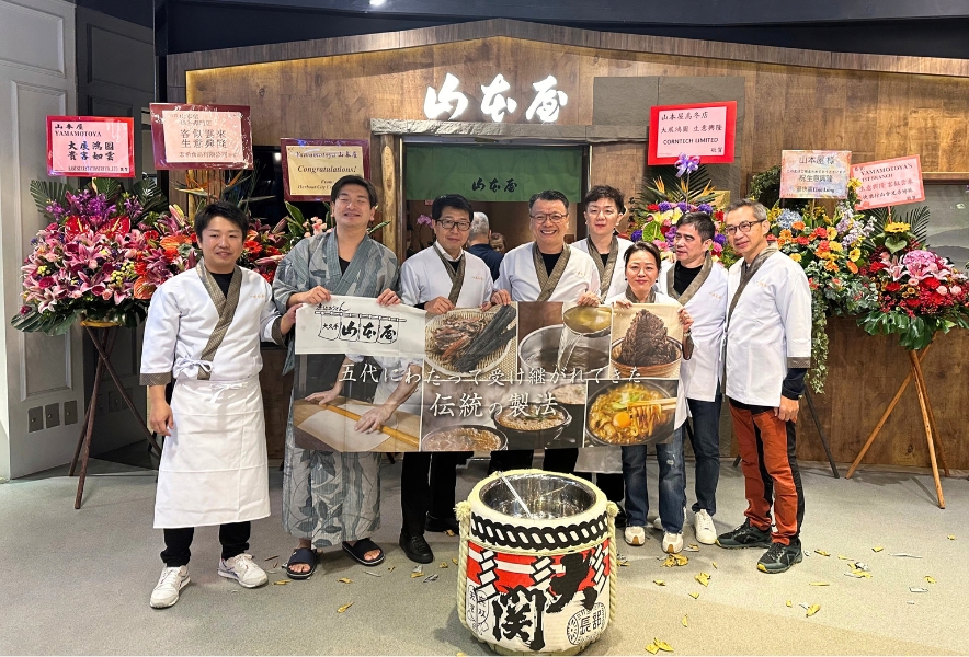 香港の山本屋大久手1号店オープン時の集合写真。祝い花と鏡開きの樽の前に立つ5代目兄弟と関係者